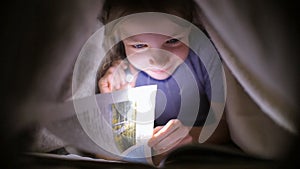 Little girl is reading a book under a blanket with a flashlight in a dark room at night