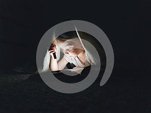 little girl is reading a book under a blanket with a flashlight in a dark room at night