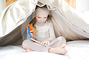 Little girl reading fairy tales book under the covers at the evening with lantern.
