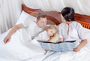 Little girl reading with her parents in bed