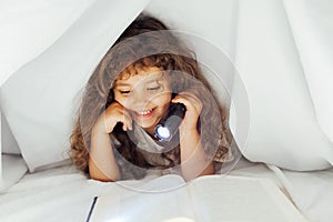 Little girl reads a book with a flashlight under a blanket in bed