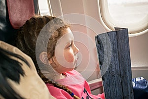 A little girl reads the menu on the plane. A child is sitting near the window of an aircraft. Baby travels by plane and reads a