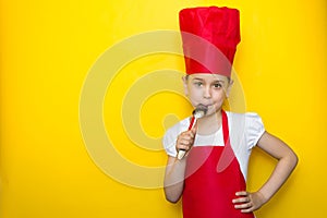 Little girl in a red chef`s suit lick the spoon, delicious taste, on yellow background with copy space