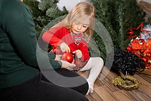 Little girl in red dress with her mother sitting on the floor in the living room among Christmas tree details and