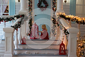 Little girl in red dress sitting on veranda of decorated house, Christmas lights, New Year`s Eve. next are the lights. decorated