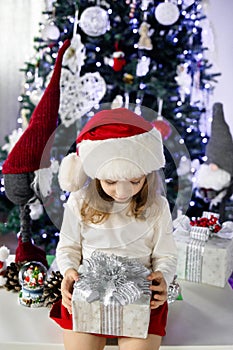 A little girl in a red Santa& x27;s Hat is sitting under the Christmas tree and playing with a gift box. Christmas and
