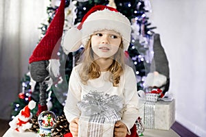 A little girl in a red Santa& x27;s Hat is sitting under the Christmas tree and playing with a gift box. Christmas and