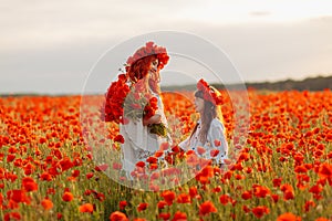 Little girl with redhead mother in white dresses and wreathes poses with bouquet of poppies at summer sunset on poppy field