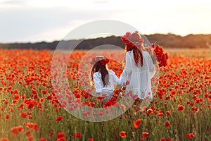 Little girl with redhead mother in white dresses and wreathes walking with bouquet of poppies on poppy field at summer sunset