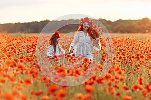 Little girl with redhead mother in white dresses and wreathes walking with bouquet of poppies on poppy field at summer sunset