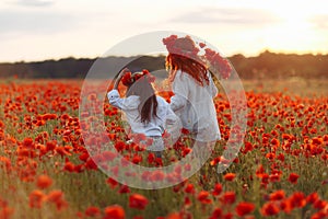 Little girl with redhead mother in white dresses and wreathes walking with bouquet of poppies on poppy field at summer sunset
