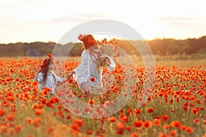 Little girl with redhead mother in white dresses and wreathes walking with bouquet of poppies on poppy field at summer sunset