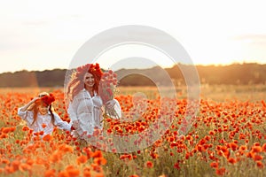 Little girl with redhead mother in white dresses and wreathes walking with bouquet of poppies on poppy field at summer sunset