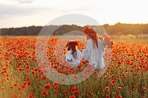 Little girl with redhead mother in white dresses and wreathes walking with bouquet of poppies on poppy field at summer sunset