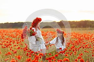 Little girl with redhead mother in white dresses and wreathes walking with bouquet of poppies on poppy field at summer sunset