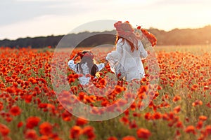 Little girl with redhead mother in white dresses and wreathes walking on poppy field with bouquet of poppies at summer sunset