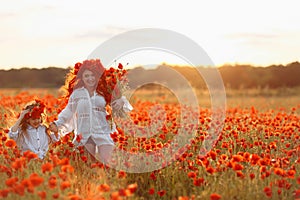 Little girl with redhead mother in white dresses and wreathes walking on poppy field with bouquet of poppies at summer sunset