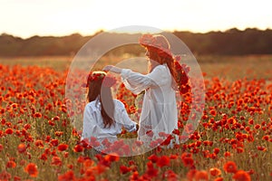 Little girl with redhead mother in white dresses and wreathes walking on poppy field with bouquet of poppies at summer sunset
