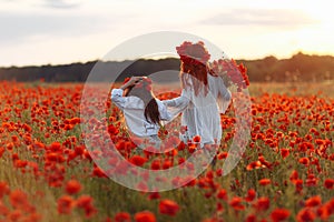 Little girl with redhead mother in white dresses and wreathes walking on poppy field with bouquet of poppies at summer sunset