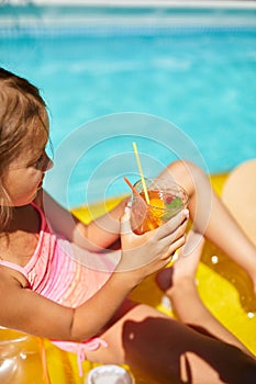 Little girl relaxing in swimming pool, enjoying suntans, drink a juice on inflatable yellow mattress
