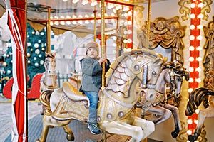 Little girl rides a toy horse on a carousel in the square near a decorated Christmas tree