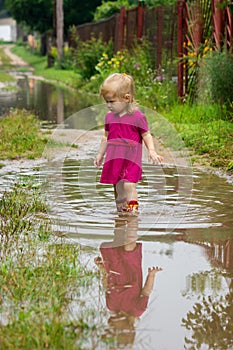 Little girl in rubber boots stands in the middle of a dirty large deep puddle on a country sandy road after rain