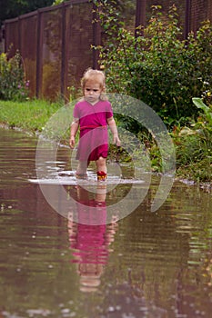 Little girl in rubber boots stands in the middle of a dirty large deep puddle on a country sandy road after rain