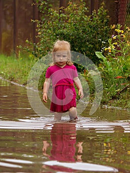 Little girl in rubber boots stands in the middle of a dirty large deep puddle on a country sandy road after rain