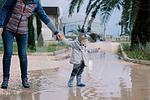 Little girl in rubber boots stands in a puddle holding her mother hand. Cropped