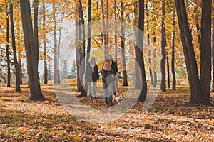 Little girl running with her dog jack russell terrier among autumn leaves. Mother and grandmother walks behind