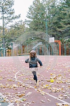 Little girl runs along a basketball court strewn with fallen leaves in the park. Back view