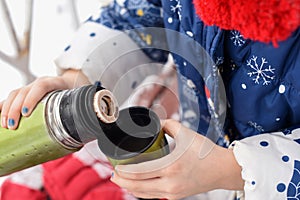 A little girl`s hands pourring tea from a thermos to a cup againts a scenic winter landscape