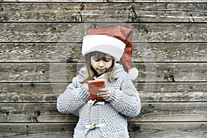 Little girl in Santa Claus hat having fun with a red cellphone outdoors during Christmas time - Little girl standing against a
