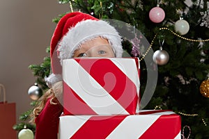 A little girl in a Santa Claus hat is hiding behind a stack of New Year`s boxes with gifts.