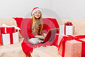 Little girl in Santa Claus hat sitting on sofa at home with laptop  with Christmas gifts