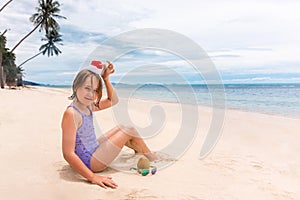 Little girl in Santa Claus red hat plays with coconut on sand on a tropical beach. Holiday tropic summer concept