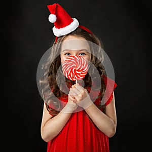 Little girl in santa hat with candy, isolated dark background.