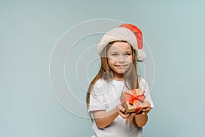 Little girl in Santa hat and with Christmas gift on blue background. Copy space. Xmas and Happy New year concept