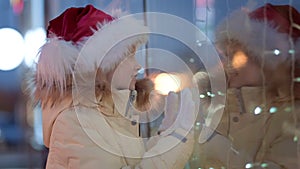 Little Girl in Santa hat with Gifts box looking and dreaming in Christmas window shopping on traditional Christmas