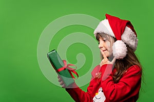 A little girl in a Santa hat holds a Christmas present in her hand and tries to guess the contents of the box. Isolated