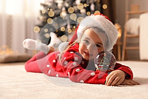 Little girl in Santa hat playing with snow globe on floor