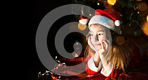 Little girl in a Santa hat and red dress under Christmas tree is dreaming, waiting for the holiday, lying on a plaid blanket. A