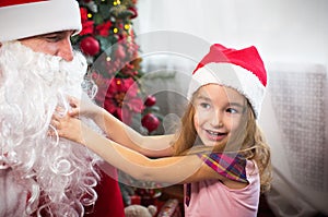 Little girl in Santa hat on Santa`s lap near Christmas tree in Christmas decor. Shows a box with a gift, a soft toy, touches his b
