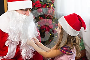Little girl in Santa hat on Santa`s lap near Christmas tree in Christmas decor. Shows a box with a gift, a soft toy, touches his b