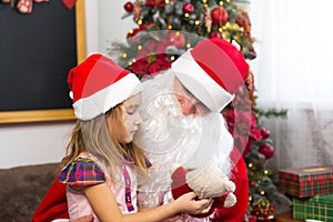 Little girl in Santa hat on Santa`s lap near Christmas tree in Christmas decor. Shows a box with a gift, a soft toy, touches his b