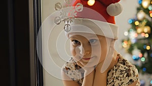 Little girl in Santa hat sitting in the evening in the room by the window