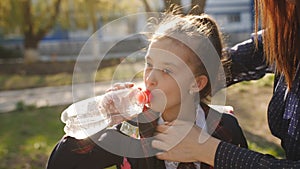 Little girl in school uniform drinks water from a plastic bottle. Mother adjusts her daughter tie, and the girl quenches