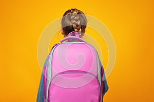 Little girl schoolgirl stands with her back on a yellow background. The child holds a satchel.