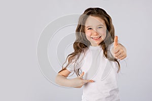 Little girl showing thumbs up gesture in a white T-shirt on white background.