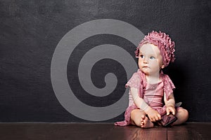 Little girl siting 1 year on a brown wooden floor.  The girl looks aside, holding phone in hand. Thoughtful look. Copy space
